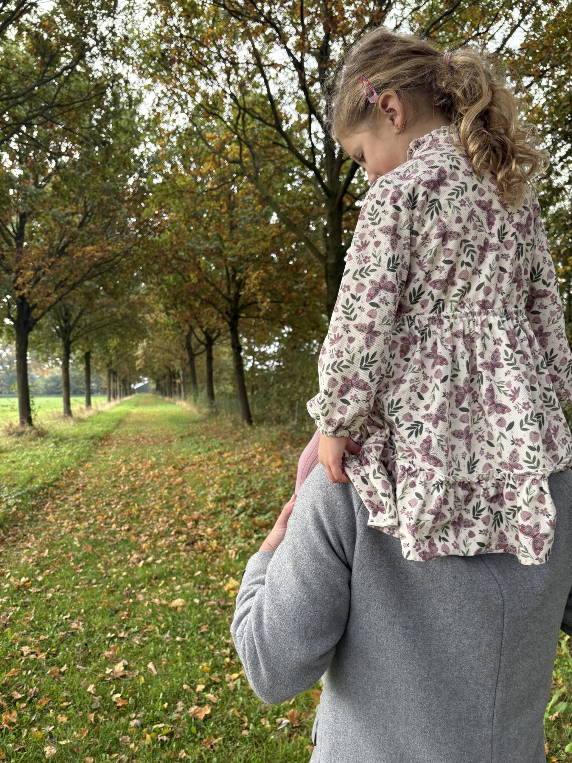Een close-up vanaf de achterkant van een volwassene in een grijze jas die een jong kind op de schouders draagt. Het kind draagt een jurk met een bloemen- en vlinderpatroon. Ze bevinden zich op een zandpad omzoomd door bomen met herfstbladeren.