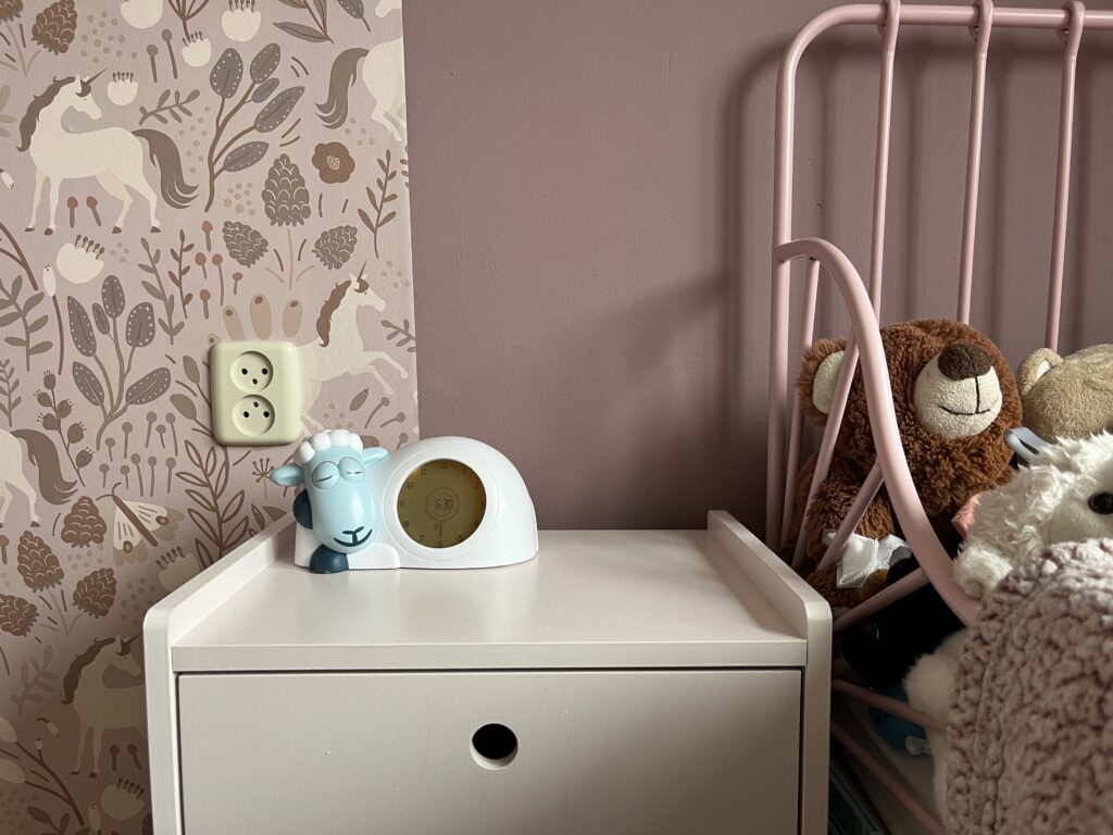 Een wit nachtkastje in een kinderkamer met daarop een witte slaaptrainer in de vorm van een schaap met gesloten ogen. Op de achtergrond roze muren en behang met eenhoorns. Naast het kastje staat een roze spijlenbed met een bruine teddybeer.